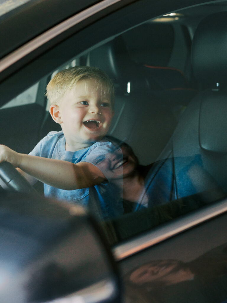 toddler son smiling at mom from inside the driver seat of her car while she smiles back in the reflection of the car window