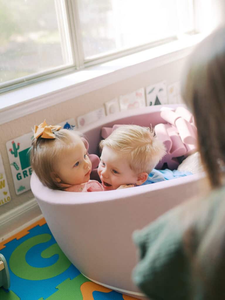 siblings playing in a foam pit in a bright living room with mom watching over them