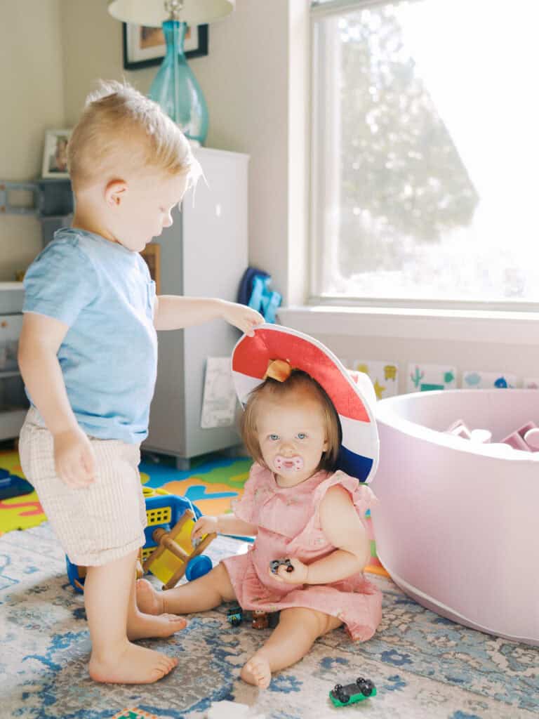 toddler boy in a bright room placing a cowboy hat on baby sister